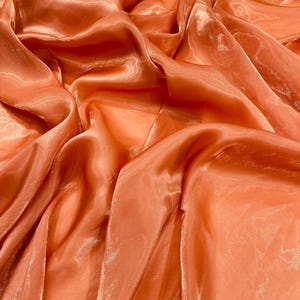 May include: Close-up of flowing, peach-colored fabric with a glossy sheen. The fabric is draped and folded, creating shadows and highlights that emphasize its texture. The material appears lightweight and delicate, with a subtle shimmer.