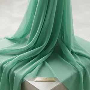 May include: A length of jade-colored stretch power mesh fabric draped in soft folds. A small rectangular label at the base displays the text: JADE 58/60" WIDE SOLID STRETCH POWER MESH. The material features a fine, semi-sheer, grid-like texture.