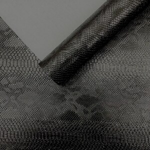 May include: A close-up shot of black faux leather with a snakeskin pattern. The material is partially rolled, revealing the textured surface and the underside of the material. The lighting creates highlights and shadows, emphasizing the scales.