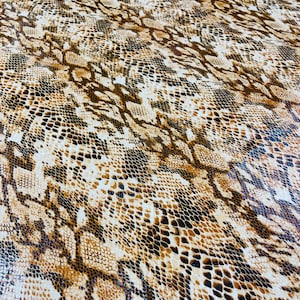 Pu&ograve; includere: Un primo piano di un tessuto con motivo a pelle di serpente marrone e bianco. Il tessuto ha una superficie testurizzata e un motivo ripetitivo di squame marroni, bianche e nere.