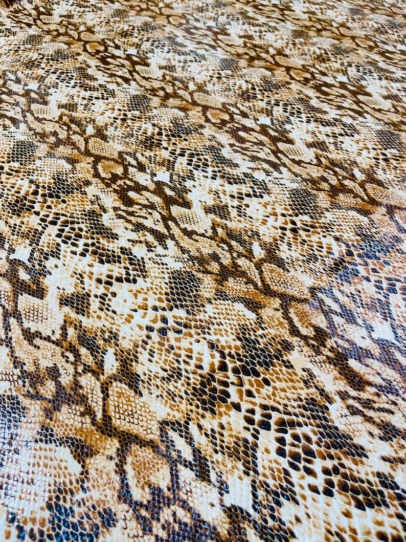 Pu&ograve; includere: Un primo piano di un tessuto con motivo a pelle di serpente marrone e bianco. Il tessuto ha una superficie testurizzata e un motivo ripetitivo di squame marroni, bianche e nere.