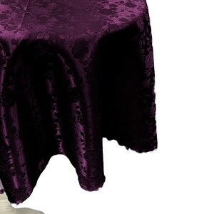 May include: A round, deep purple tablecloth with a floral pattern. The fabric drapes over a white table base. The tablecloth is made of a shiny, textured material, and the floral design is visible throughout the fabric.