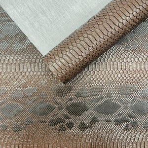 May include: Close-up of brown faux snakeskin fabric, partially rolled, with a textured pattern. The material has a metallic sheen and a reptile scale design. A section of gray fabric is visible in the background.