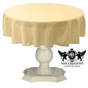 May include: A round, pale yellow tablecloth draped over a white pedestal table with a decorative base. The tablecloth is smooth and wrinkle-free. The table is round and has a pedestal base with a decorative design.