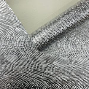 May include: A roll of silver faux leather material with a reptile scale pattern. The material is shiny and reflective, with a textured surface. The material is partially unrolled, revealing the underside.