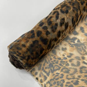 May include: Rolled fabric with a leopard print pattern. The fabric features a tan background with dark brown and black spots. The material is rolled up, revealing the edge of the fabric.