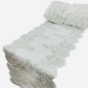 Może przedstawiać: Biała koronka z delikatnym wzorem kwiatowym i koralikowymi akcentami. Koronka ma falowany brzeg i idealnie nadaje się do dodania elegancji każdemu projektowi.