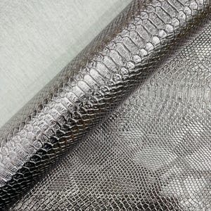 May include: A close-up of a roll of silver faux leather fabric with a snakeskin pattern. The fabric has a metallic sheen and a textured surface, with a light gray background.