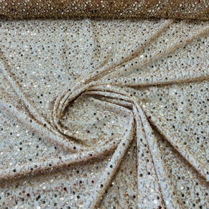 May include: Close-up of a cream-colored fabric covered in gold sequins. The fabric is draped and folded, with a roll of the same sequined material visible in the background. The sequins catch the light, creating a shimmering effect.