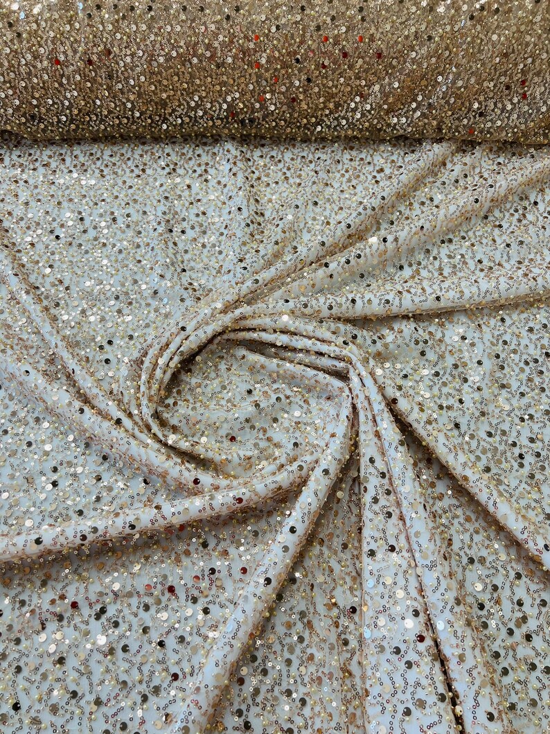 May include: Close-up of a cream-colored fabric covered in gold sequins. The fabric is draped and folded, with a roll of the same sequined material visible in the background. The sequins catch the light, creating a shimmering effect.