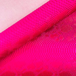 May include: Vibrant pink snakeskin leather with a textured, scaled pattern. The leather is partially rolled, revealing a lighter pink fabric underneath. The bright colour and unique texture make it suitable for various crafting projects.