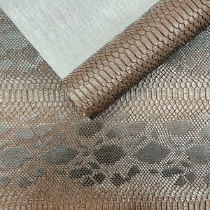 May include: A roll of brown faux leather fabric featuring a realistic snakeskin texture with a repeating diamond pattern. The material has a glossy finish and is displayed against a light grey, fine-textured background surface.