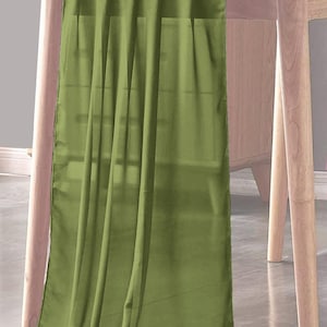 Può includere: Runner da tavolo verde oliva drappeggiato su un tavolo in legno chiaro. Il tessuto è liscio e fluido, con pieghe morbide. Il runner da tavolo aggiunge un tocco di colore ed eleganza al tavolo.
