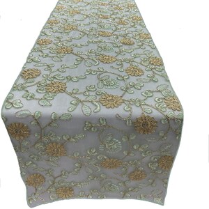 May include: A light green table runner with a floral pattern in gold and green. The runner is made of a sheer fabric and has a delicate, embroidered design.
