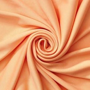 Könnte beinhalten: Ein einfarbiger, hellpfirsichfarbener Stoff mit einer glatten, matten Textur. Das Material ist in einem zentralen Spiralmuster angeordnet, wodurch tiefe, weiche Falten und Schatten entstehen, die vom Mittelpunkt des Stoffes ausgehen.