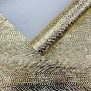 May include: Gold faux leather fabric with a snakeskin texture. The material is shown flat and rolled, revealing a detailed pattern and a metallic sheen. The background is a soft white.