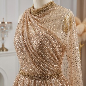 May include: A champagne-colored formal gown with a high neckline and long, sheer sleeves. The dress is embellished with sequins and a beaded belt. The bodice features a draped design, and the neckline is adorned with small beads.