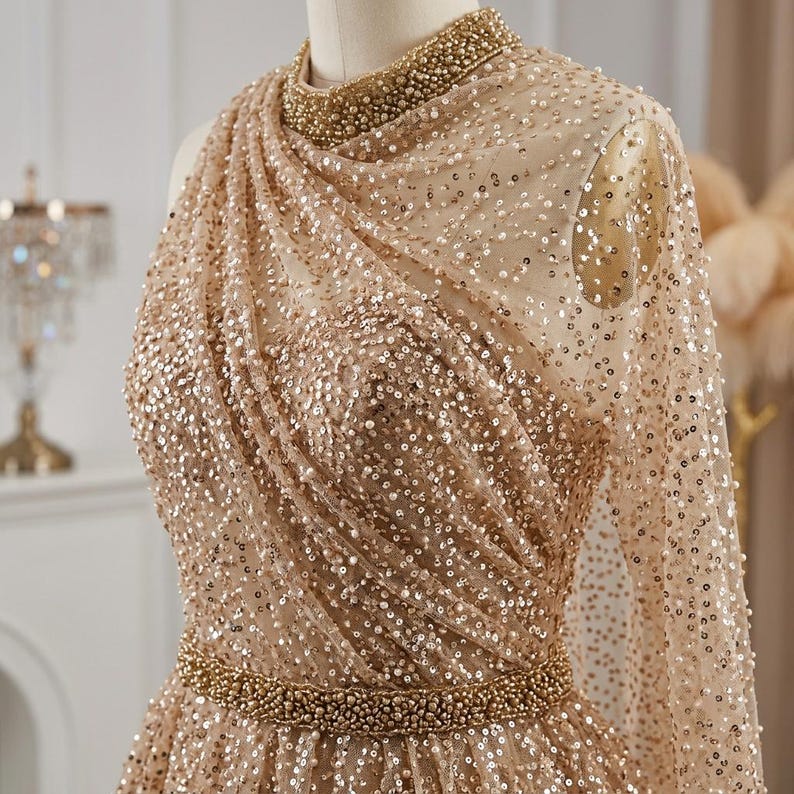 May include: A champagne-colored formal gown with a high neckline and long, sheer sleeves. The dress is embellished with sequins and a beaded belt. The bodice features a draped design, and the neckline is adorned with small beads.