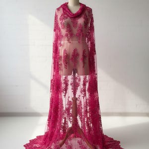 May include: A floor-length fuchsia lace cape featuring intricate floral embroidery and delicate beadwork throughout the sheer mesh fabric. The garment has a draped cowl neckline and a scalloped hemline that pools on the floor around the base.