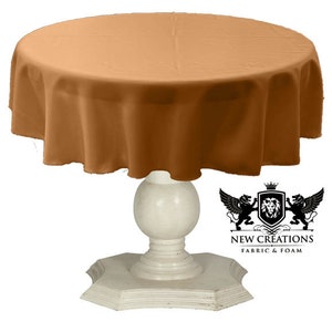 May include: A round, brown tablecloth draped over a white pedestal table. The tablecloth is made of a smooth, silky fabric. The table has a round top and a pedestal base with a decorative, fluted design. The text "NEW CREATIONS FABRIC & FOAM" is visible in the lower right corner of the image.