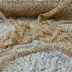 May include: Close-up of a gold sequin fabric. The fabric is draped, showing a dense pattern of small, reflective sequins. The sequins are in various shades of gold, creating a shimmering effect. The fabric is likely used for apparel or decorative purposes.