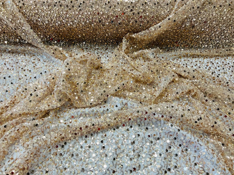 May include: Close-up of a gold sequin fabric. The fabric is draped, showing a dense pattern of small, reflective sequins. The sequins are in various shades of gold, creating a shimmering effect. The fabric is likely used for apparel or decorative purposes.