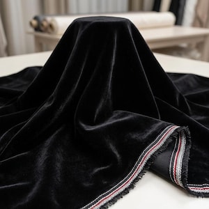 May include: A close-up of black velvet fabric draped over a form. The fabric has a soft, luxurious texture. The edge of the fabric is visible, trimmed with a striped pattern in red, white, and black.