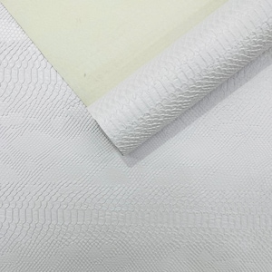 Peut inclure: Une feuille de matériau blanc avec un motif texturé en peau de serpent, partiellement enroulée pour révéler un envers lisse beige clair. La surface présente un motif d'écailles gaufré répétitif qui recouvre toute la partie blanche visible.