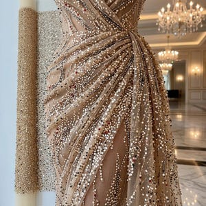 May include: A beige evening gown with intricate sequin and bead embellishments. The dress features a draped design and a flowing skirt. Rolls of matching fabric are visible in the background, suggesting custom tailoring.