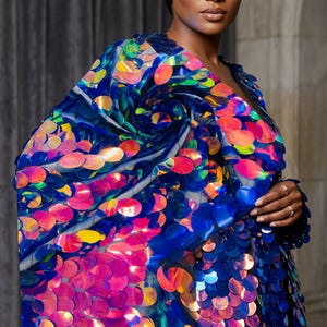 Könnte beinhalten: Blaue Paillettenjacke mit V-Ausschnitt. Die Jacke ist mit großen, schillernden Pailletten in Blau, Rosa, Orange und Gelb verziert. Die Pailletten bilden ein wirbelndes Muster auf der Schulter. Ein auffälliges Kleidungsstück.