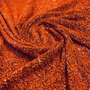 May include: Close-up of a fabric with a textured surface made of small, orange sequins. The sequins are tightly packed together, creating a shimmering effect.