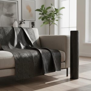 May include: A dark charcoal gray throw blanket with a textured snakeskin-like pattern draped over a light beige sofa, accompanied by a matching cylindrical roll of the same dark gray material standing upright on the floor beside the furniture.