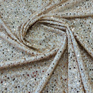 May include: Close-up of a cream-colored fabric covered in small, shimmering gold sequins. The fabric is draped and folded, creating a textured effect. The sequins catch the light, adding a touch of glamour and sparkle to the material.