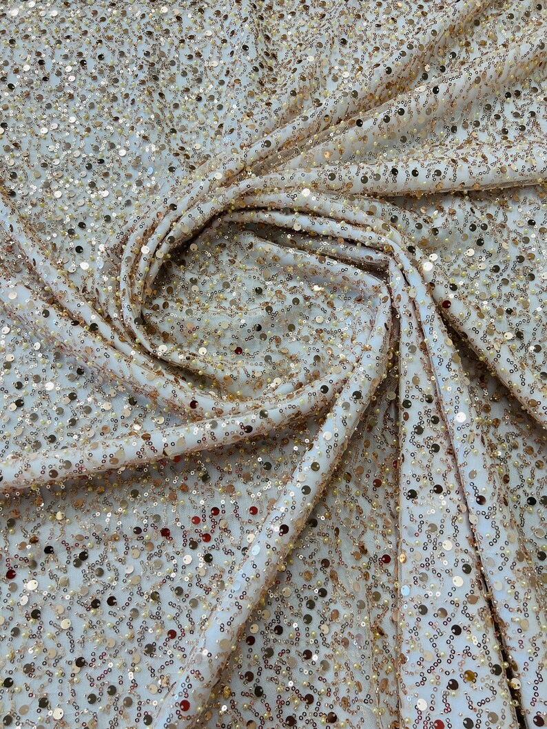 May include: Close-up of a cream-colored fabric covered in small, shimmering gold sequins. The fabric is draped and folded, creating a textured effect. The sequins catch the light, adding a touch of glamour and sparkle to the material.