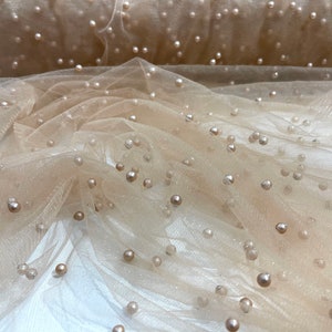 Puede incluir: Tela de tul beige con adornos de perlas dispersos. La tela tiene una textura suave y delicada y un brillo sutil.