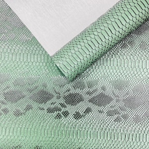 May include: A sheet of mint green material featuring a textured snakeskin pattern with dark grey diamond-shaped markings. The material is rolled at the top, revealing a smooth, light grey underside with a subtle, fine-grained fabric texture.