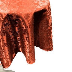 以下が含まれることがあります： 花柄の丸いオレンジ色のテーブルクロス。テーブルクロスは柔らかく絹のような生地でできており、上品な光沢があります。