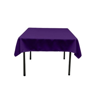 May include: A square, deep purple tablecloth draped over a black-legged table. The fabric has a smooth texture, and the corners of the cloth hang down. The table is set against a white background.