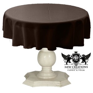May include: A brown round tablecloth draped over a white pedestal table. The tablecloth is made of a smooth, wrinkle-resistant fabric. The table has a round top and a pedestal base with a decorative design.