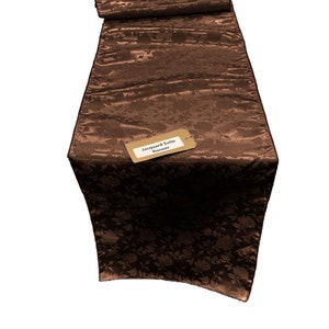 May include: A brown jacquard satin table runner with a floral pattern. The runner has a tag that says "Jacquard Satin Runner".
