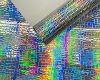 Op de afbeelding: Een rol en een vel iriserend imitatieleer met een krokodillenleertextuur. Het materiaal reflecteert een regenboog aan kleuren, waaronder blauw, groen, geel en paars, waardoor een glinsterend effect ontstaat. Geschikt voor knutselen en doe-het-zelf projecten.