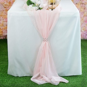 Puede incluir: Un camino de mesa rosa claro con un adorno de cristal, drapeado sobre un mantel blanco. El camino de mesa está hecho de tela transparente y es perfecto para agregar un toque de elegancia a cualquier mesa.