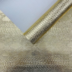 May include: A sheet of metallic gold material featuring a raised, repeating snakeskin texture. The material is partially rolled over itself, revealing a smooth, light-coloured underside against the textured, reflective gold surface.