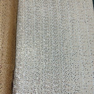 May include: A roll of shimmering fabric with a dense pattern of small, reflective sequins. The fabric is a warm gold color, with a slight texture visible. The sequins catch the light, creating a sparkling effect. Ideal for apparel and craft projects.