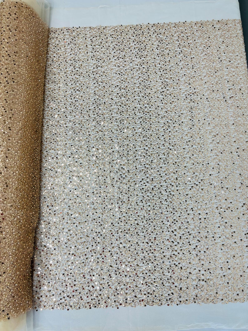 May include: A roll of shimmering fabric with a dense pattern of small, reflective sequins. The fabric is a warm gold color, with a slight texture visible. The sequins catch the light, creating a sparkling effect. Ideal for apparel and craft projects.