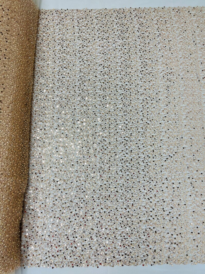 May include: A roll of gold sequin fabric. The fabric is covered in small, round sequins that catch the light. The sequins are a mix of gold and silver, creating a shimmering effect. The fabric is likely used for apparel or decorative purposes.