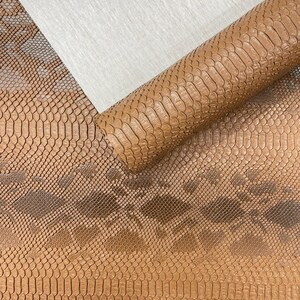 May include: A close-up of brown faux snakeskin fabric. The fabric is rolled up in a cylinder shape, with a portion of the material unrolled, revealing the detailed scale pattern. A piece of white fabric is in the background.