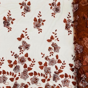 Puede incluir: Tela de encaje floral marrón con flores 3D. La tela tiene un fondo de malla delicado y presenta flores en relieve texturizadas de color marrón claro.