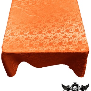 Puede incluir: Mantel de mesa naranja con estampado floral. El mantel está hecho de una tela satinada y tiene un brillo sutil. El mantel está drapeado sobre una mesa y los bordes cuelgan.
