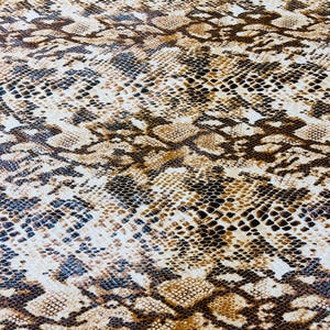 Pu&ograve; includere: Un primo piano di un tessuto con motivo a pelle di serpente marrone e bianco. Il tessuto ha una superficie testurizzata e un motivo a scaglie ripetuto.